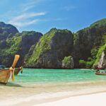 Traditional long-tail boats resting on the shore of a turquoise beach surrounded by towering cliffs. One of the top reasons to visit Thailand is its picturesque beaches, making Thailand a great holiday destination for beach lovers.