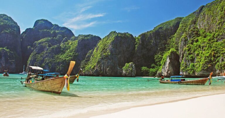 Traditional long-tail boats resting on the shore of a turquoise beach surrounded by towering cliffs. One of the top reasons to visit Thailand is its picturesque beaches, making Thailand a great holiday destination for beach lovers.