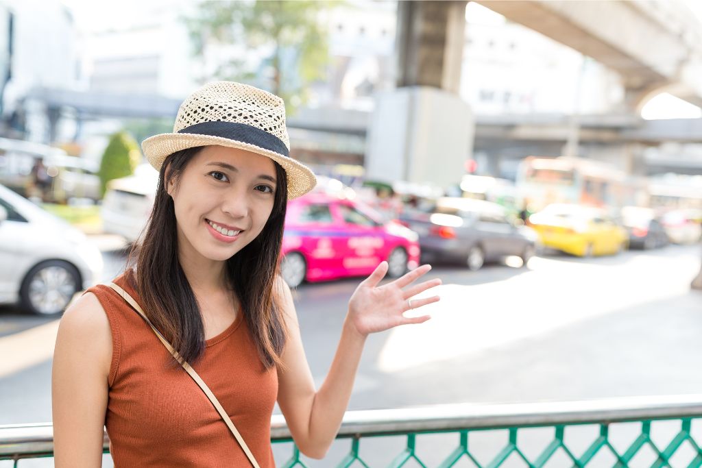 A smiling woman waves on a busy Bangkok street, with colorful taxis in the background. One reason to visit Thailand is its vibrant city life, making Thailand a nice place to explore.