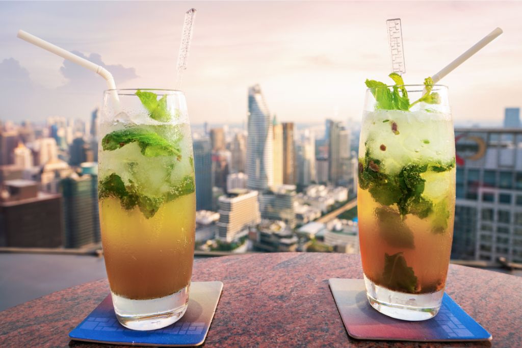 two mojito cocktails with fresh mint served on a rooftop bar in Bangkok with a view of the city skyline. Reasons to go to Thailand include the stunning city views and unique experiences like rooftop dining.