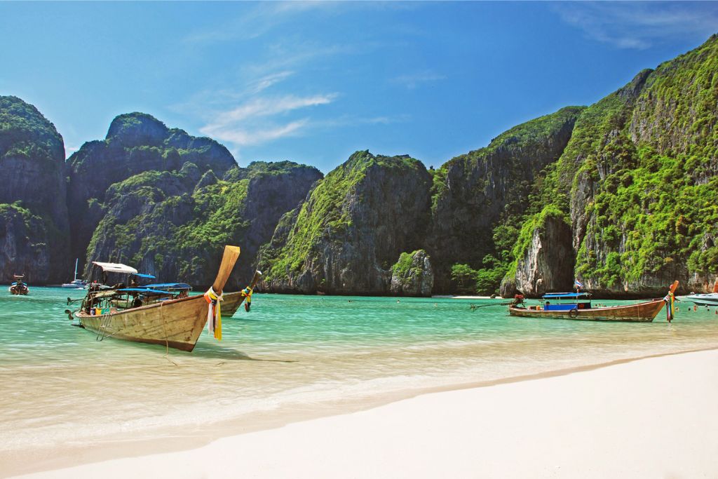 A traditional Thai long-tail boat anchored on a pristine beach with emerald green water, surrounded by dramatic cliffs. Why visit Thailand? Its beautiful beaches make it a great holiday destination.