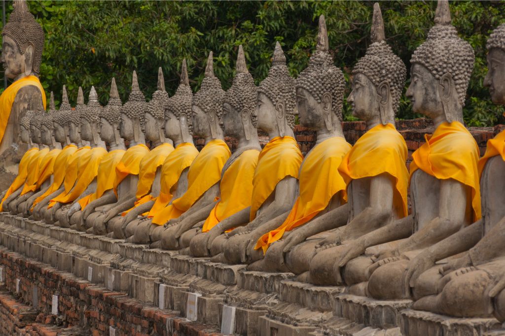 A row of seated Buddha statues draped in yellow robes at a Thai temple. Visiting Thailand is worth it for the serene spiritual sites, which are key reasons why people go to Thailand.