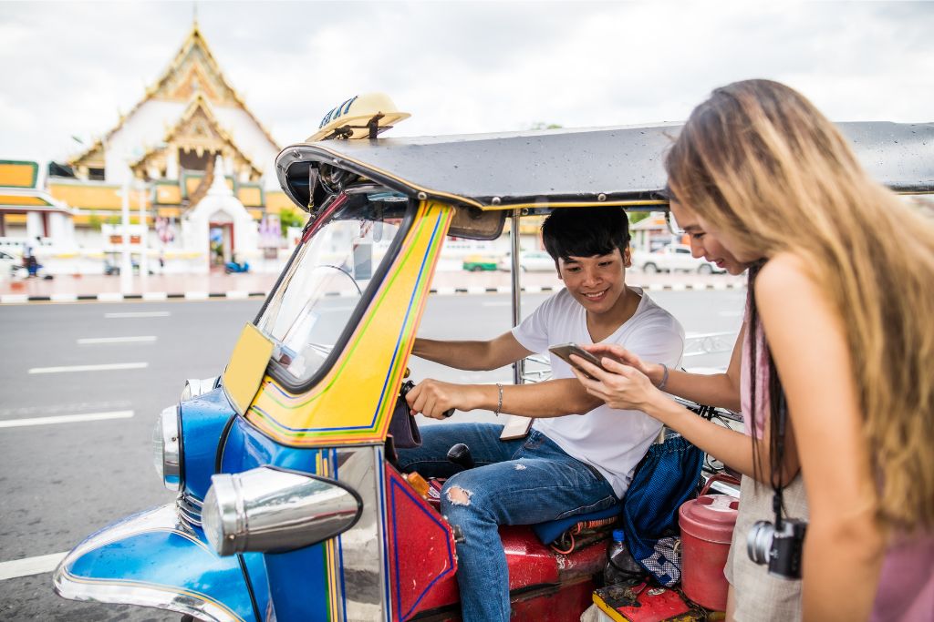 A tourist shows something on their phone to a smiling tuk-tuk driver in Bangkok. Why go to Thailand? Exploring the streets in a tuk-tuk is one of the reasons you should visit Thailand.