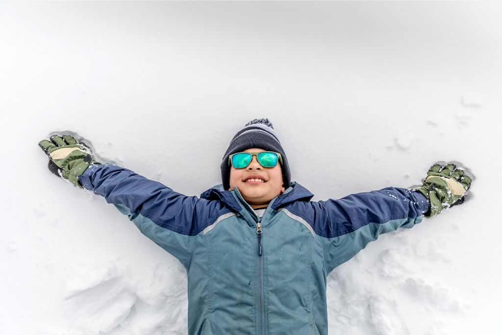 A smiling boy wearing winter gear and sunglasses lying on his back in the snow, arms spread wide as he makes a snow angel. Perfect for enjoying snowy holidays in Singapore Snow City.