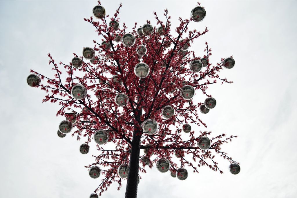 A striking red artificial tree adorned with reflective globe ornaments, set against a cloudy sky. A beautiful winter wonderland aesthetic for holiday destinations in Singapore.