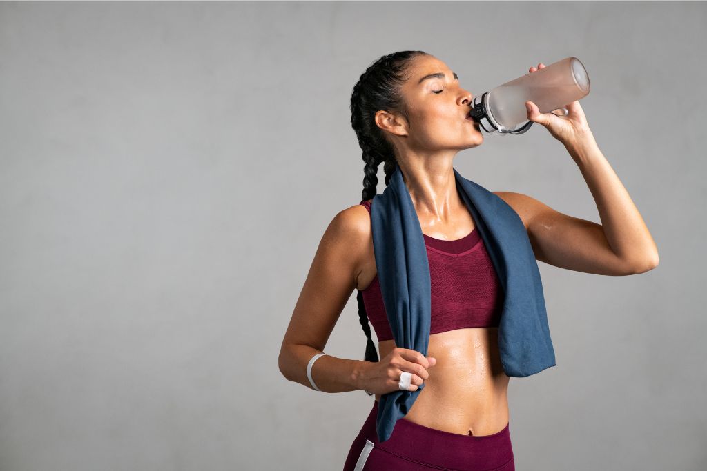  A fit woman in workout gear drinking water from a bottle, her skin glistening after exercise. Represents staying active and healthy during winter travels in Singapore.