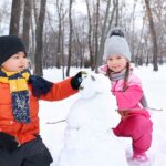 Two children in colorful winter outfits building a snowman in a snowy park, surrounded by leafless trees. A cozy and joyful winter activity, evoking a winter wonderland aesthetic.