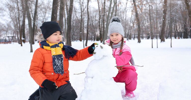 Two children in colorful winter outfits building a snowman in a snowy park, surrounded by leafless trees. A cozy and joyful winter activity, evoking a winter wonderland aesthetic.