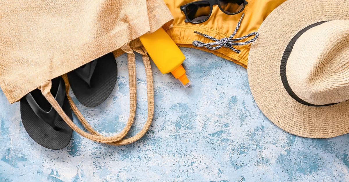 A flat lay of summer essentials displays a straw tote spilling out beach must-haves like black flip-flops, sunscreen, sunglasses, and a beach hat. These beach bag essentials are perfect for a well-prepared beach day.