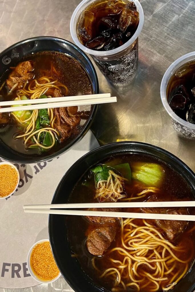 Overhead shot of two bowls of Taiwanese beef noodle soup served with vegetables and chili oil, alongside iced drinks on a metal table. Sampling flavorful Asian food like this is one of the best things to do in Singapore for food lovers and night market explorers.