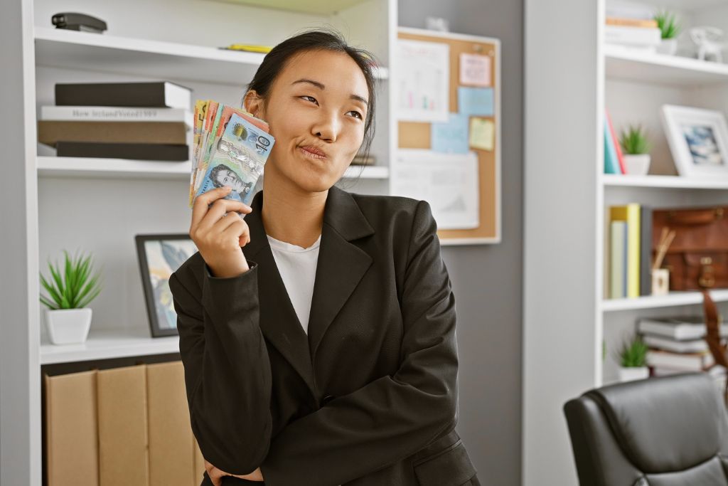 Young woman in a black blazer playfully holding Australian dollar bills in an office, visually representing financial independence—a persuasive factor in how to convince your parents to let you travel alone or go on a solo trip.