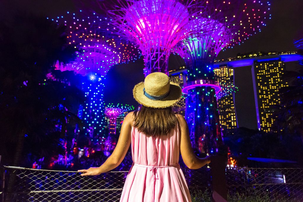 A woman in a straw hat gazes at the brightly lit Supertree Grove and Marina Bay Sands during the Gardens by the Bay light show. Garden Rhapsody is one of the most memorable night attractions and free light shows in Singapore.