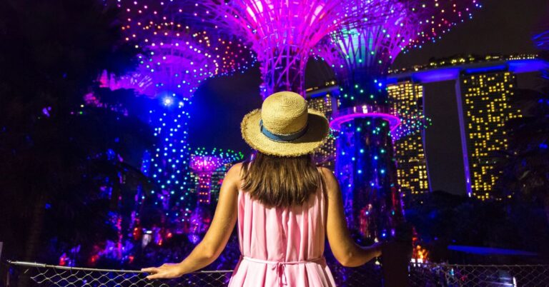 A woman in a pink dress and straw hat stands under glowing Supertrees at night, watching the Gardens by the Bay light show with Marina Bay Sands illuminated in the background. This moment captures the immersive beauty of Garden Rhapsody, a top light show in Singapore.