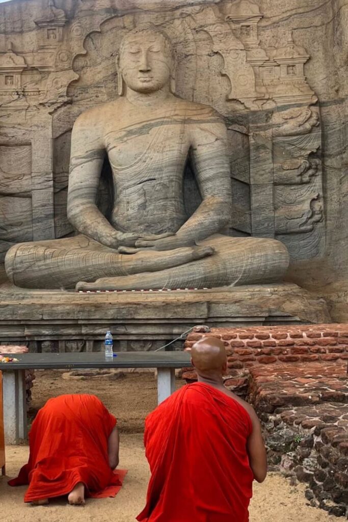 wo monks in red robes kneeling before a large Buddha statue carved into stone in Sri Lanka, reflecting spiritual solo journeys and offering a serene example of solo travel tips for introverts or mindful female travelers.