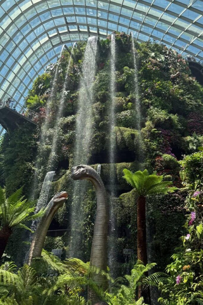 Lush indoor waterfall inside the Cloud Forest dome at Gardens by the Bay, with mist falling over ferns and life-size dinosaur sculptures. This indoor attraction is a top thing to do in Singapore alongside the Garden Rhapsody light show.