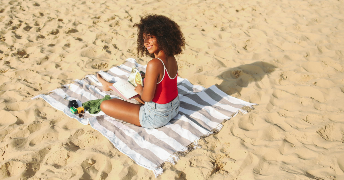 A woman in a red swimsuit and denim shorts sits on a striped beach blanket, smiling while reading a book. A camera, sunglasses, and other small items are beside her on the sand. This peaceful moment highlights beach hacks for solo female travelers, such as bringing a waterproof beach bag for valuables to keep personal items safe.