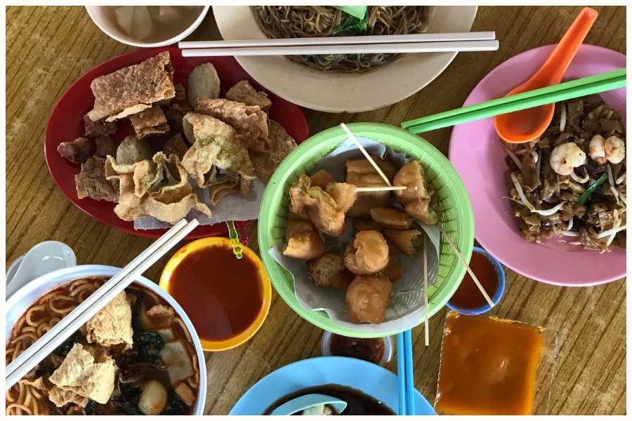 A variety of hawker food dishes arranged on a table, including fried snacks, noodles, and dipping sauces. The image captures the diverse street food to try in Singapore, showcasing what to eat in Singapore's best hawker centers.