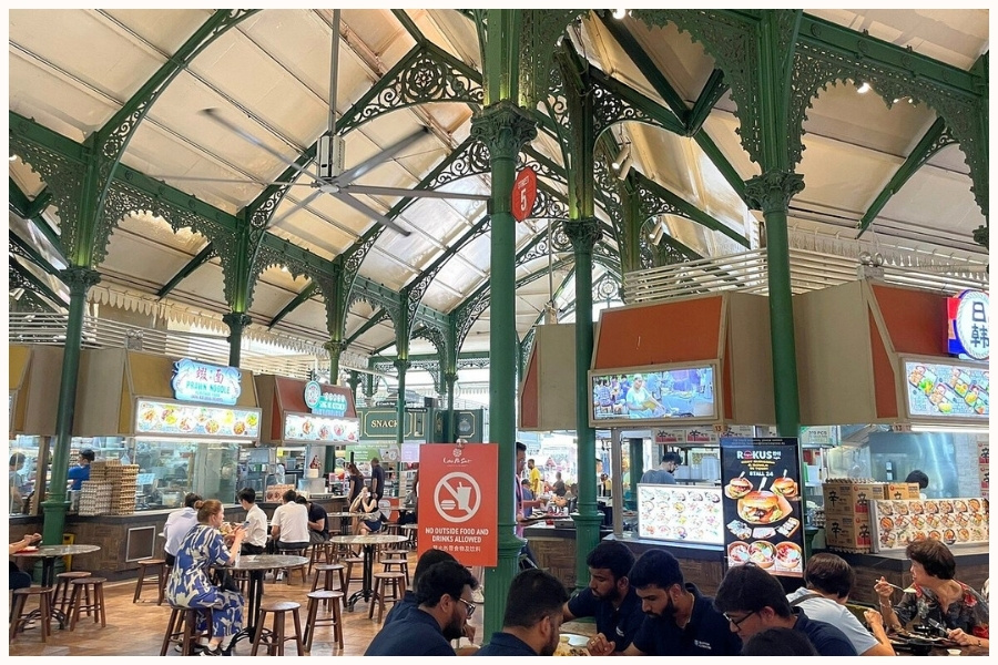 A bustling hawker center with ornate green pillars, multiple food stalls, and people enjoying their meals. This scene showcases one of the top hawker centers in Singapore, offering a variety of street food to try in Singapore.