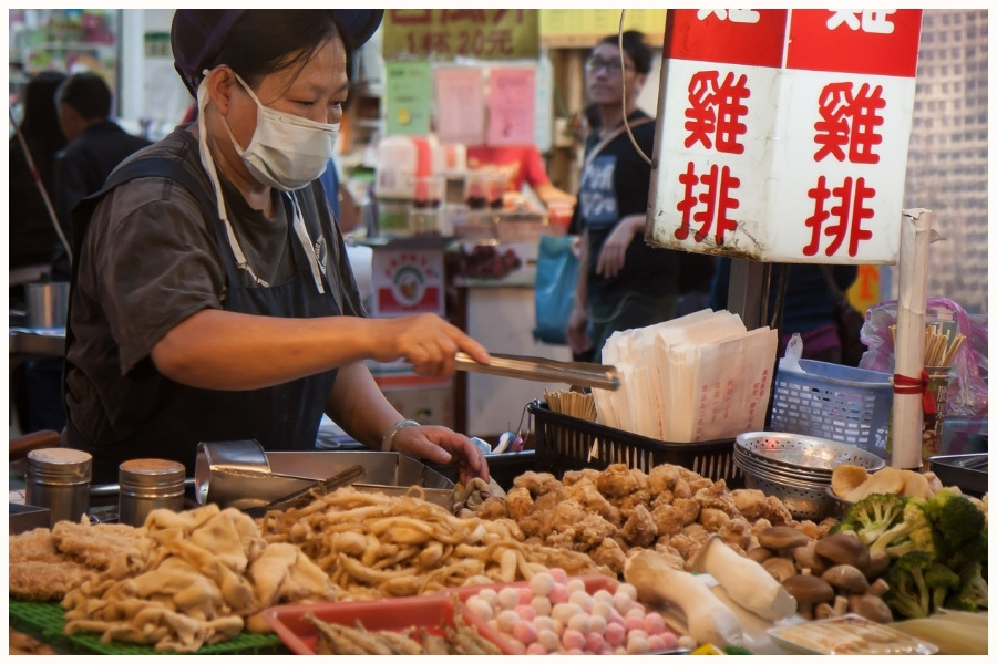 A hawker stall vendor wearing a mask, serving a variety of fried and skewered items. This image captures the essence of Singapore food markets and showcases some of the best street food in Singapore.