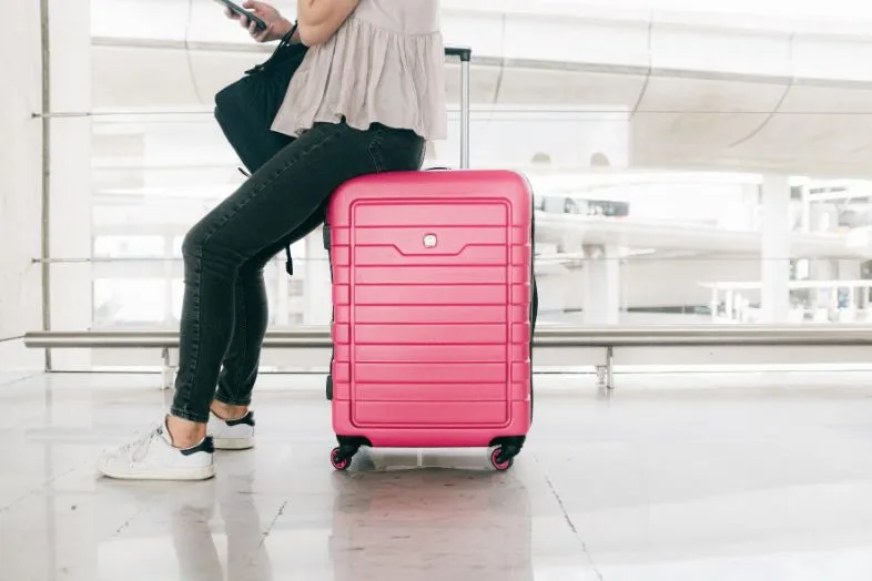 A traveler sits on a bright pink suitcase at the airport, making their luggage stand out easily among common black and brown bags. Using vibrant colors like pink is a fun way to make luggage noticeable and easy to identify."