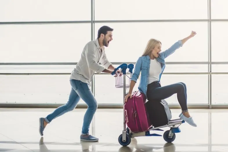 A man pushes a woman sitting on an airport luggage cart, having fun while traveling. Decorating your luggage with bright colors and unique luggage covers is a playful way to make your suitcase stand out at the airport and ensure it's easily recognizable