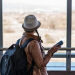 Woman wearing a backpack and holding a phone, standing at an airport window, possibly preparing for her first solo flight. A guide to essential airport procedures for first time flyers.