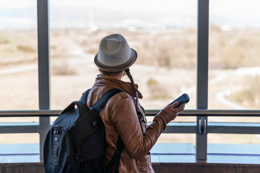 Woman wearing a backpack and holding a phone, standing at an airport window, possibly preparing for her first solo flight. A guide to essential airport procedures for first time flyers.