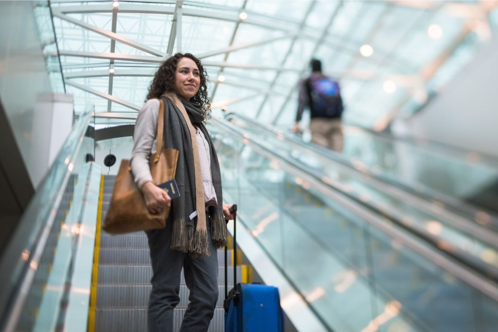 Woman standing on an escalator with luggage, holding a passport and looking ahead. A guide to airport tips for first-time flyers, focusing on how to navigate the airport as a solo traveler.