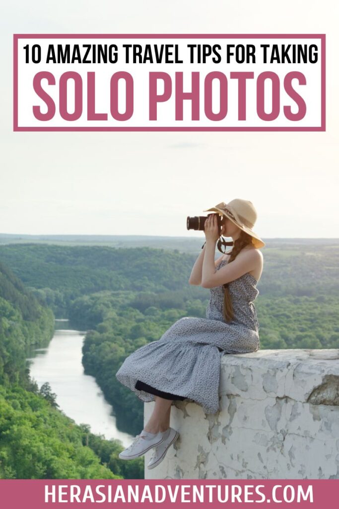 A solo traveler sits on a rock ledge overlooking a lush green valley, holding a camera and taking solo travel photos, with a guide on how to take your own pictures while traveling alone.