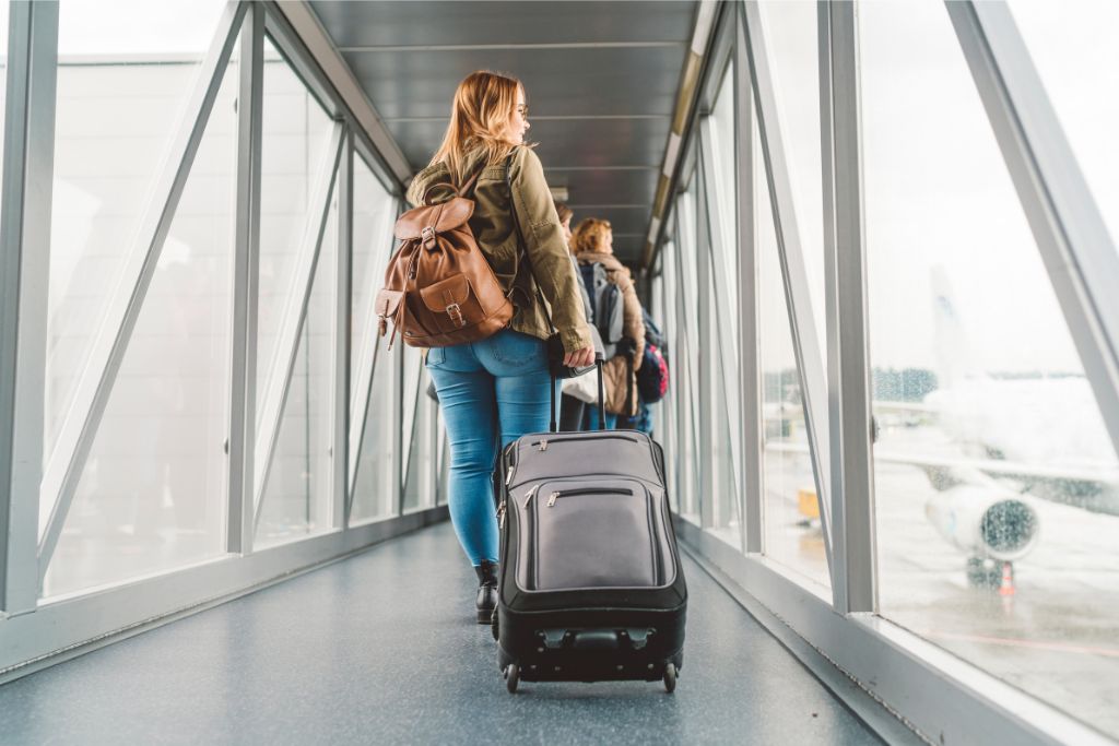 Woman walking down a glass jet bridge with a suitcase, heading towards an airplane. Ideal for first time flight travel tips, showing what to expect when boarding your first plane.