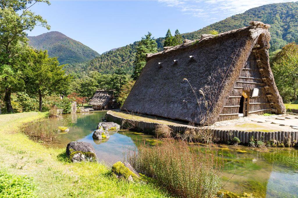 Traditional Japanese thatched houses set against a serene pond and surrounded by lush green hills, with hints of autumn colors. A peaceful and scenic fall travel destination in Japan, ideal for cultural exploration and nature photography.