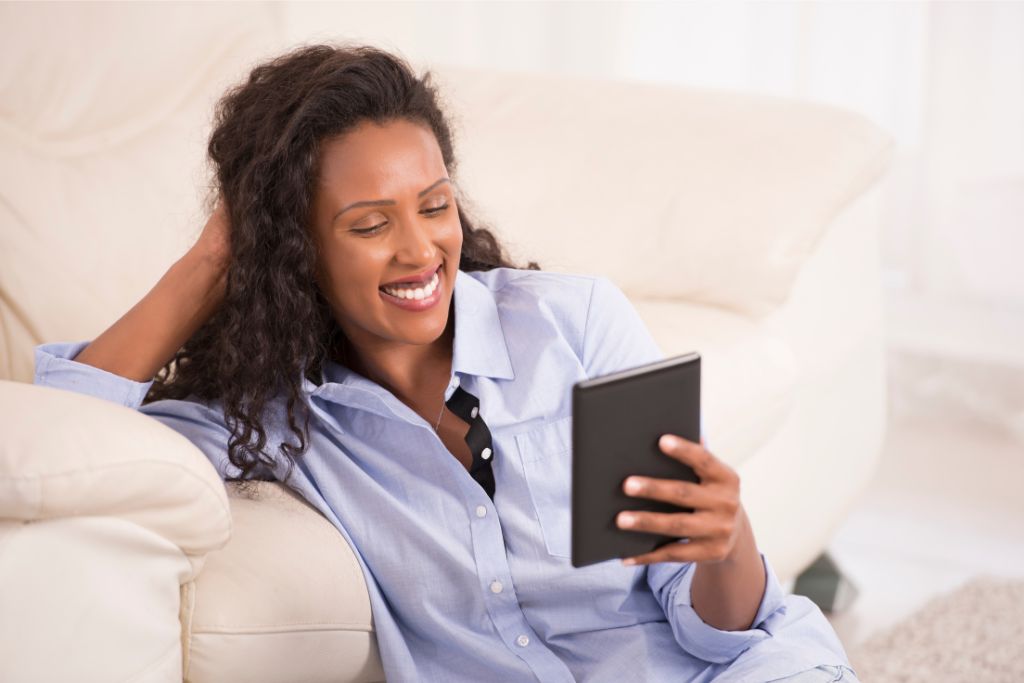 woman sitting comfortably on a couch, smiling while reading on a tablet. This image promotes relaxation and suggests the importance of packing light to avoid overpacking, ensuring you have room for digital entertainment without burdening yourself with excess luggage.