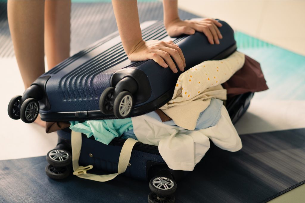 Someone struggling to close an overstuffed suitcase, a clear example of overpacking. The image emphasizes how not to overpack and offers a visual reminder of what not to pack for a trip, encouraging travelers to pack only essentials.