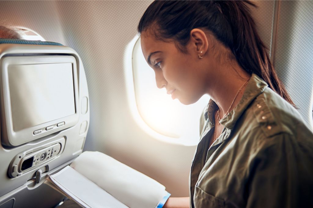 Woman reading a book inside an airplane seat, with the airplane window glowing from sunlight. Tips for first-time flyers on how to stay calm and entertained during your first flight journey.