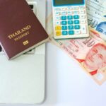 A Thai passport next to a calculator, Thai baht, and Singaporean dollars on a laptop, representing financial planning and money management while traveling in Southeast Asia. This highlights the importance of budgeting and managing multiple Asian currencies in the region.