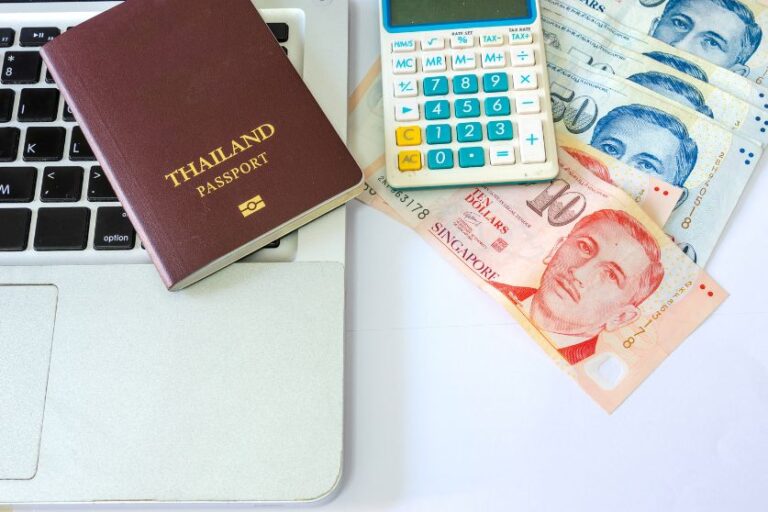 A Thai passport next to a calculator, Thai baht, and Singaporean dollars on a laptop, representing financial planning and money management while traveling in Southeast Asia. This highlights the importance of budgeting and managing multiple Asian currencies in the region.