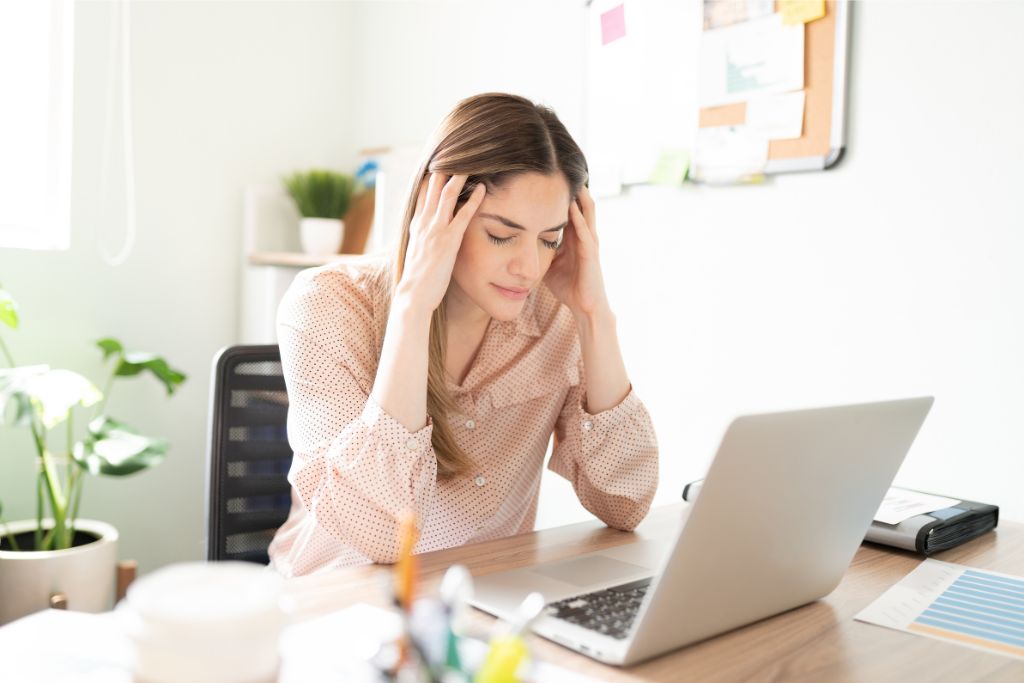 A stressed woman holding her head while working at a laptop. This image represents the frustration that can arise from overpacking and the stress of trip planning. It subtly conveys the message of avoiding overpacking to reduce travel stress.