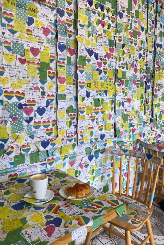 A cozy café covered in rainbow heart notes, with a latte and croissant on a table. This vibrant spot is one of the most charming autumn destinations Asia travelers can enjoy during fall holidays in Asia.