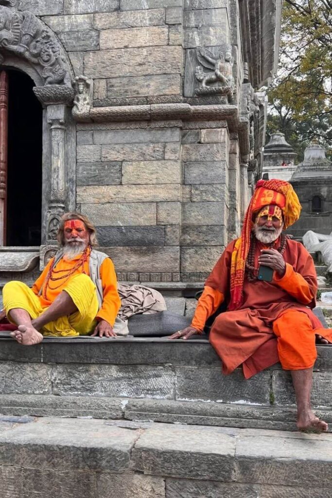 Two holy men in bright orange robes sit by a stone temple in Kathmandu. This sacred site is a fascinating Asia autumn getaway and one of the top places to visit in Asia during fall.
