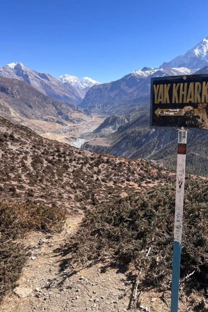 A sign for Yak Kharka overlooks rugged Himalayan peaks under a deep blue sky. This trekking spot is a top Asia place to visit in November and one of the most adventurous fall destinations in Asia.