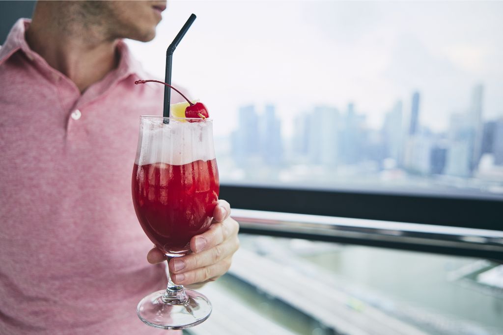 A person holds a classic Singapore Sling cocktail with a city skyline view in the background. One of the many reasons to visit Singapore is its famous cocktail culture, adding to why Singapore is worth traveling to for food and drink enthusiasts.