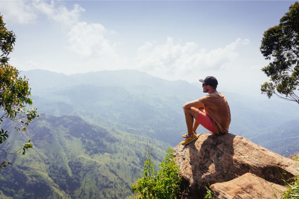 A traveler sits on a rock overlooking the lush, green mountains of Sri Lanka, embodying the adventure lifestyle. Exploring beautiful roads and hiking trails is a popular way to experience paradise while traveling Sri Lanka.