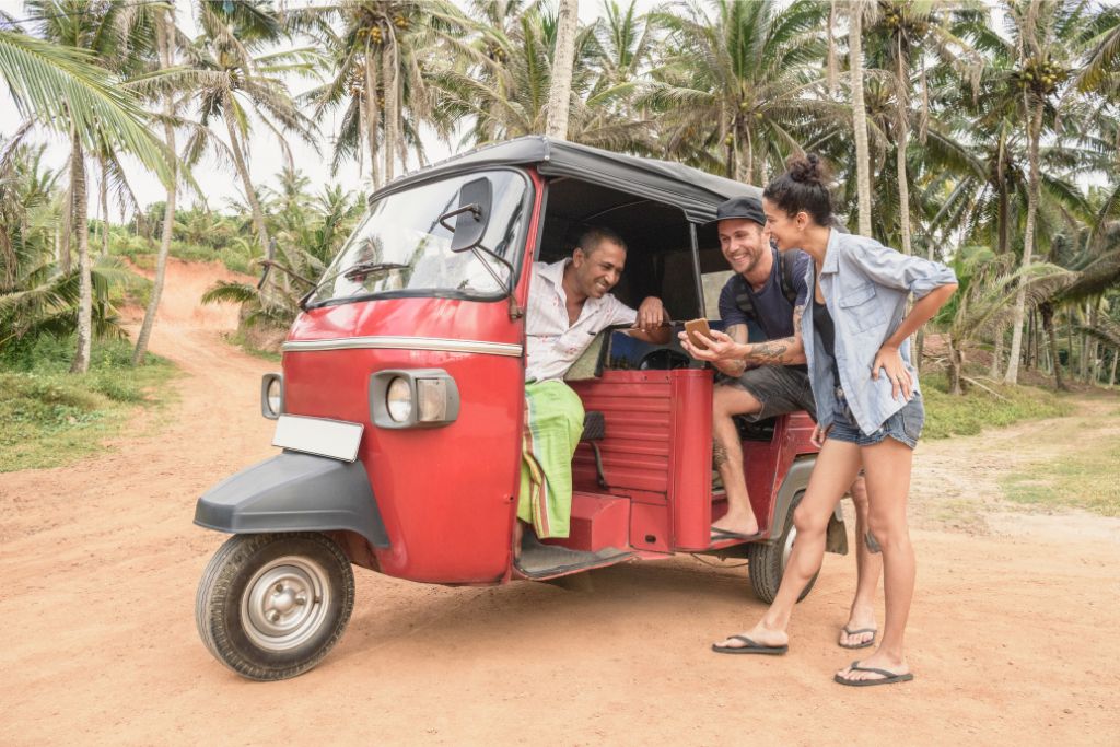 Friends chat with a local driver near a red tuk tuk in a tropical setting of palm trees in Sri Lanka. Renting a tuk tuk in Sri Lanka offers a unique and affordable way to discover the hidden beauty of this paradise island.