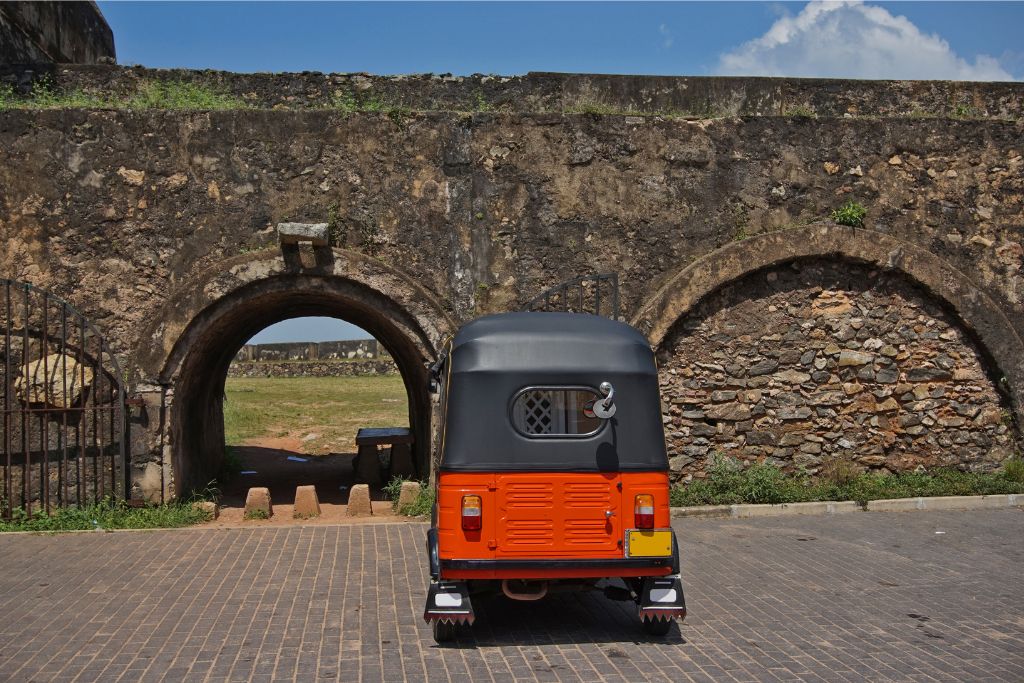 A bright orange tuk tuk parked near a historic stone arch in Sri Lanka, offering a unique way to travel and explore the country’s rich history. Tuk tuk rentals are ideal for visiting landmarks like this.