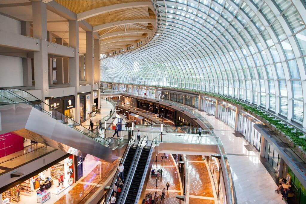 The sleek, modern interior of Marina Bay Sands shopping mall, with visitors strolling and escalators leading to high-end stores. One of the reasons to go to Singapore is its luxury shopping, demonstrating why Singapore is worth a visit for urban explorers.