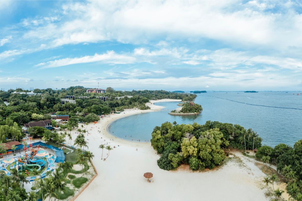 A panoramic view of a serene beach on Sentosa Island, Singapore, with clear blue skies and lush greenery. A key reason to go to Singapore is its beach destinations, offering a relaxing escape that makes Singapore worth a visit.