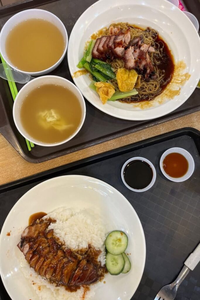 Plates of roast pork noodles and rice with soup and dipping sauces are served on food court trays, showcasing affordable and delicious Singapore hawker cuisine. Food is one of the biggest reasons why people travel to Singapore and why it's worth visiting for culinary lovers.
