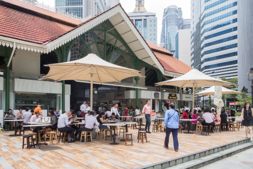 Busy lunchtime scene outside Lau Pa Sat, a historic hawker centre in Singapore’s CBD, with diners enjoying local dishes under large umbrellas amid modern skyscrapers. The vibrant street food culture here is a big reason why you should visit Singapore and one of the top culinary reasons to go to Singapore.