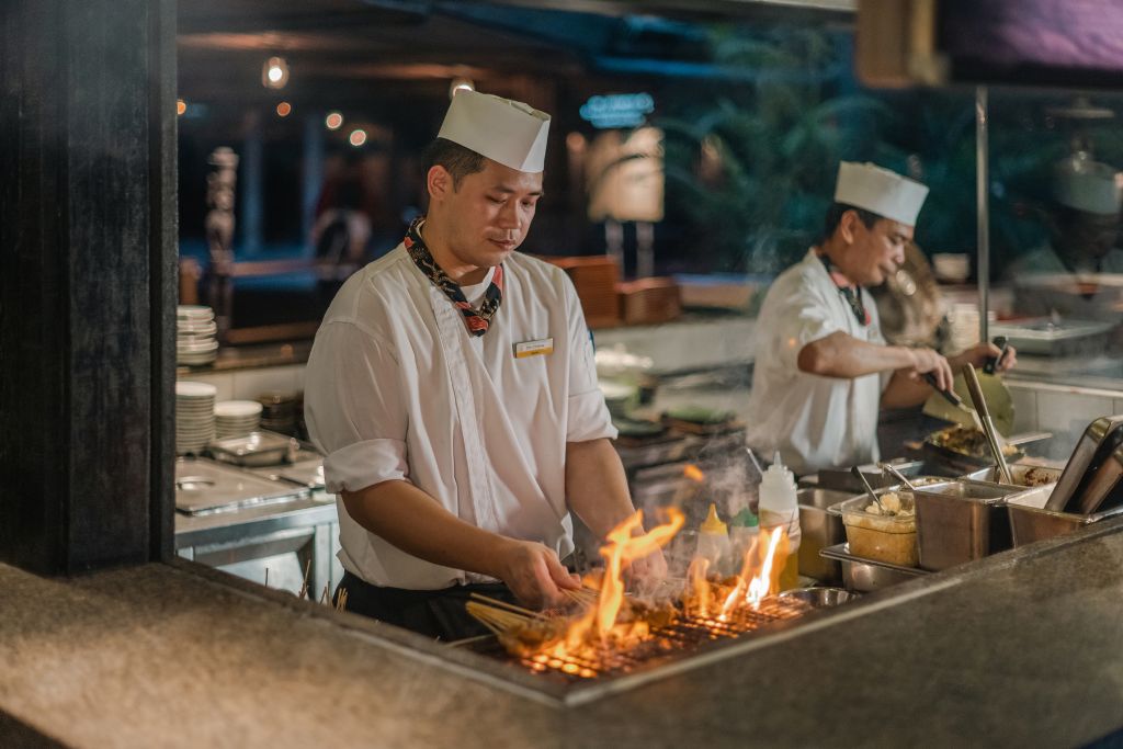 A chef grills skewered meats over an open flame at a night market-style restaurant, showcasing Singapore’s dynamic culinary scene. This fiery food experience is one of many reasons why Singapore is worth seeing for food lovers and why travel to Singapore often centers around its cuisine.