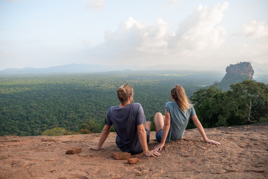 A couple sits on a rock, overlooking lush forests and landmarks like Sigiriya Rock. A great visual for Sri Lanka packing checklist and travel essentials for couples.